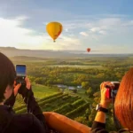 Two people in a hot air balloon basket take photos of colorful balloons floating over a lush, green landscape under a blue sky with clouds—a breathtaking adventure and one of the unforgettable things to do in Cairns. | Aussie Loop