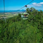 A green cable car travels above dense green forest with fields, mountains, and a blue sky in the background, offering breathtaking views along the famous Skyrail route near Kuranda Scenic Railway.